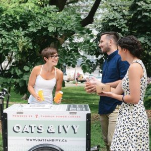 A vendor is selling juice to a couple from an Oats & Ivy branded commercial cargo vending bike in a park in Toronto Ontario