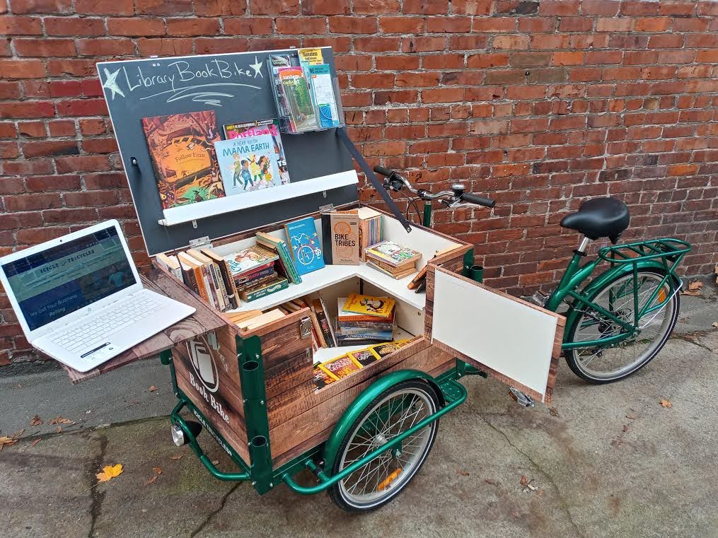 An icicle tricycles library book bike sits in front of a red brick wall. The front shelf of the trike is up and a laptop is on it. The lid is open and books sit on the shelving inside.